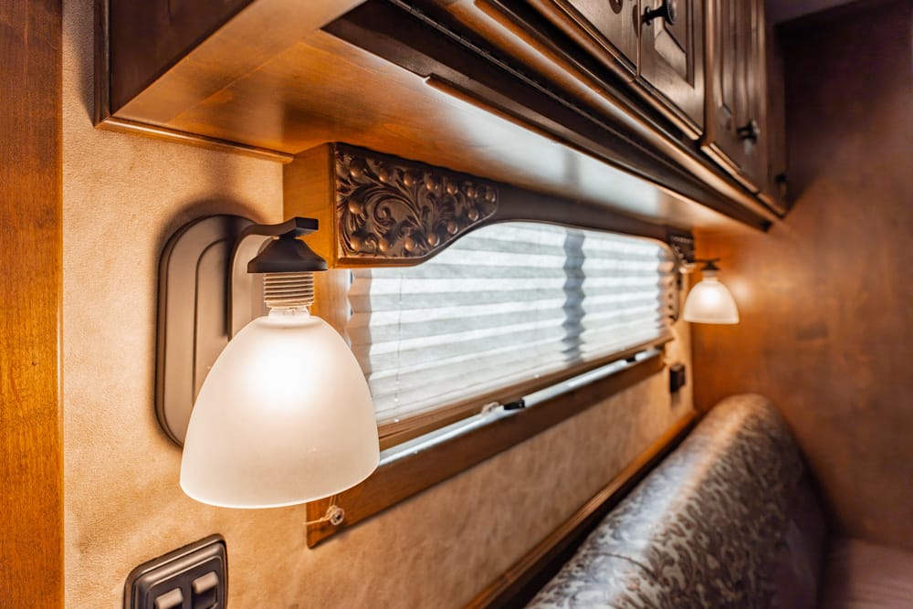 Interior of an RV with wall-mounted lamps and a pleated window shade above a patterned bench seat and wood cabinetry.
