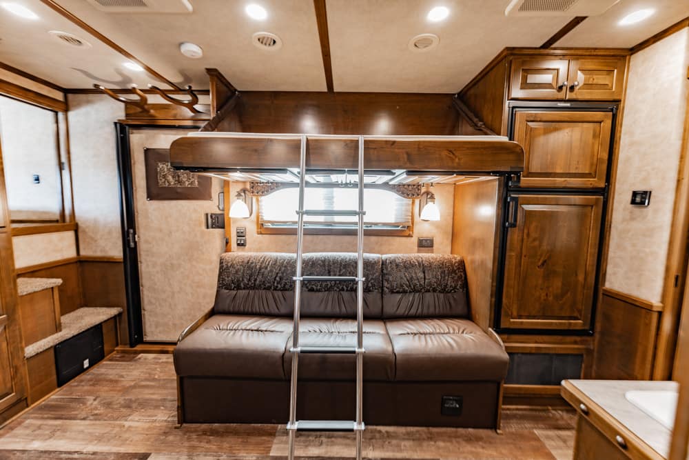 RV living area: brown leather sofa under an overhead bunk with a metal ladder, flanked by warm wood cabinets and soft lighting.