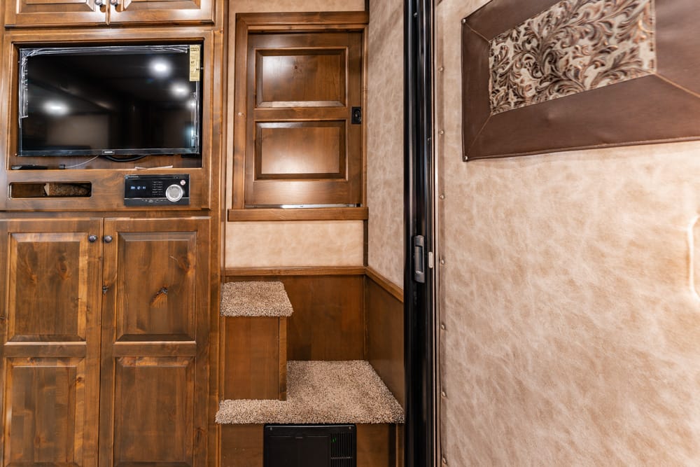 Wood-paneled RV interior with mounted TV and built-in media cabinet above carpeted steps leading to a higher deck area.