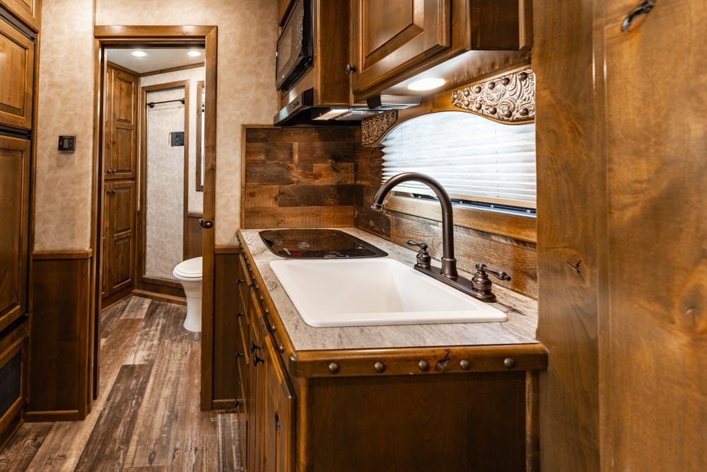 Compact RV kitchen with dark wood cabinets, white sink, black curved faucet, and stove; hallway to a small bathroom in back.