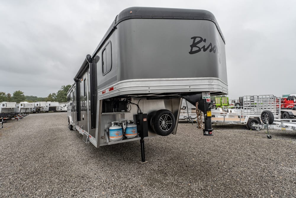 Grey fifth-wheel travel trailer parked on a gravel lot, propane tanks and a spare tire under the front overhang on a cloudy day.