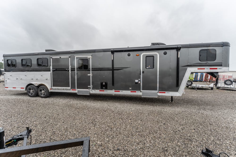 Long aluminum horse trailer with living quarters, black and silver, parked on a gravel lot under a cloudy sky.