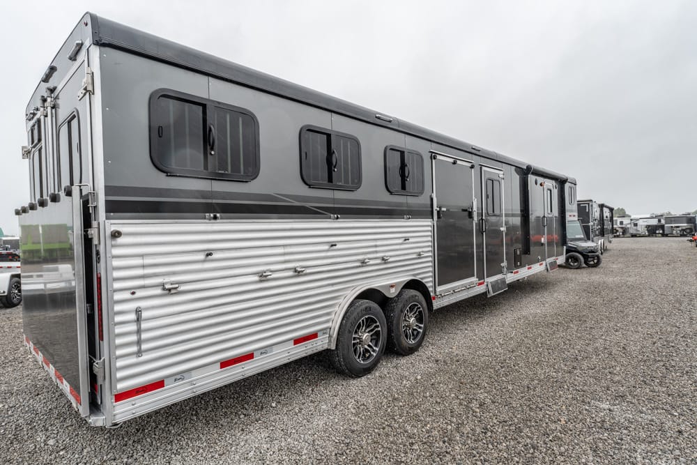 Long silver-and-gray horse trailer with black-framed windows parked on a gravel lot, other trailers visible in the background.