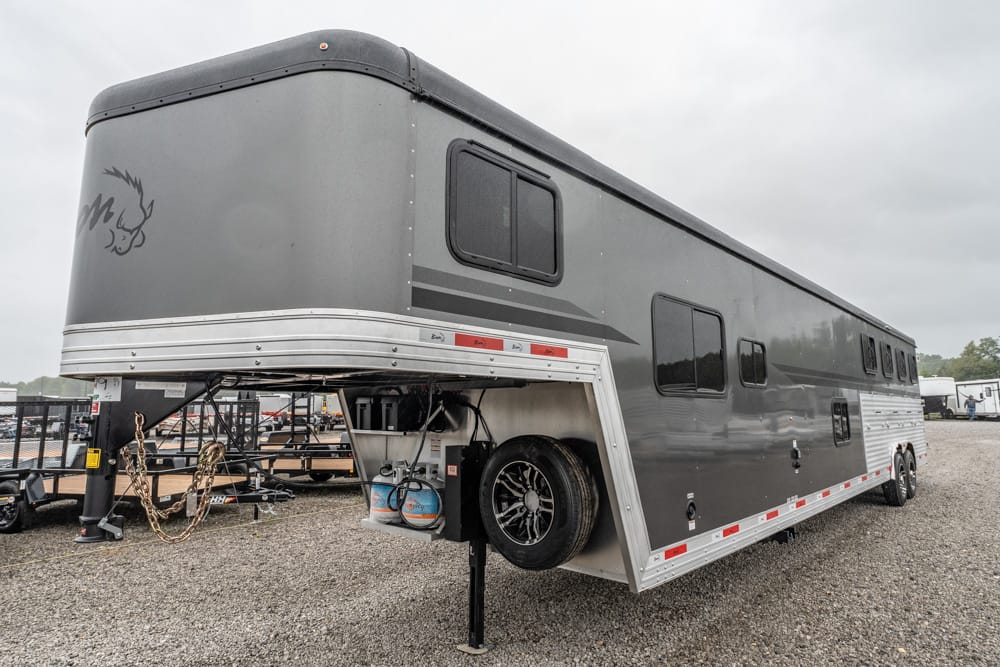 Gray, multi-window horse trailer on a gravel lot with a spare tire mounted under the front section and safety chains visible by the hitch.