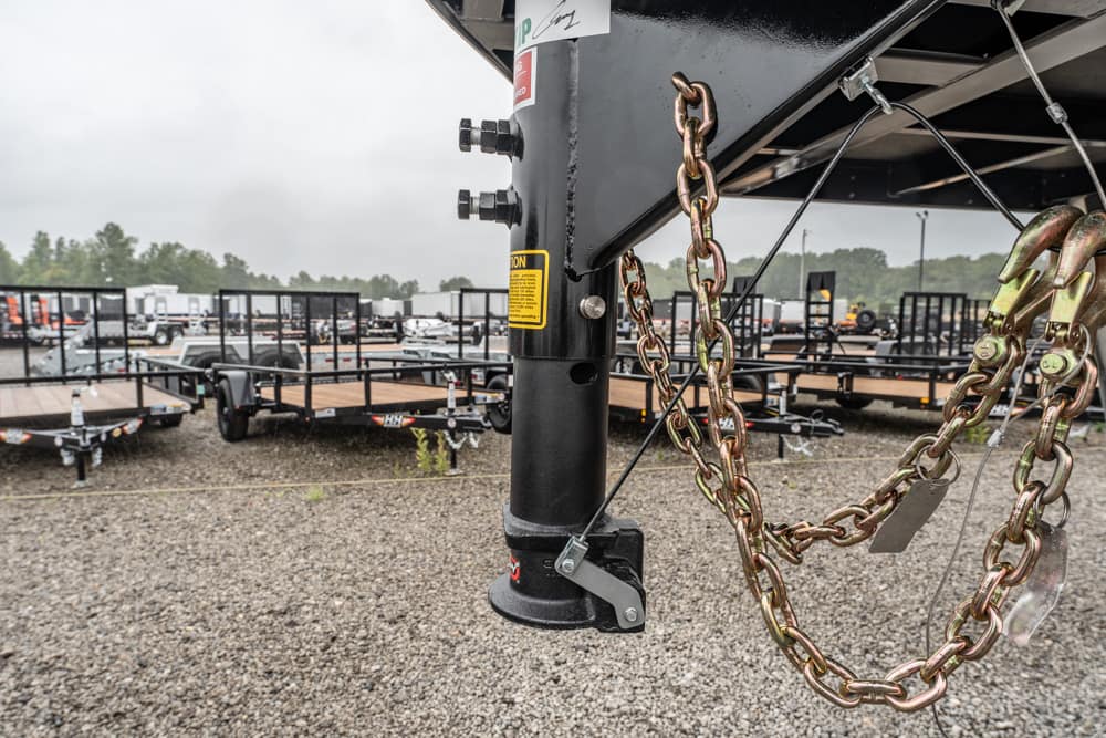 Close-up of a trailer hitch with safety chains and a latch, attached to a black vertical jack in a yard of flatbed trailers.