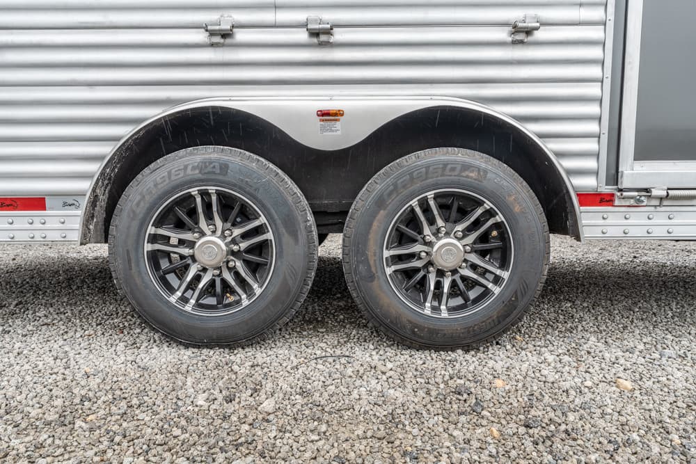 Dual trailer wheels with matching rims under a corrugated metal trailer body on a gravel surface, showing mud and scuffs.
