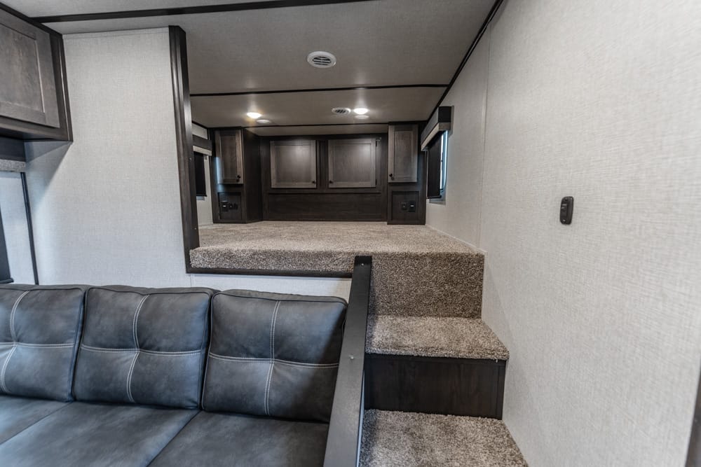 Interior of a compact RV: gray leather sofa in foreground, carpeted steps leading up to a raised kitchen/galley with dark wood cabinets and a windowed wall.