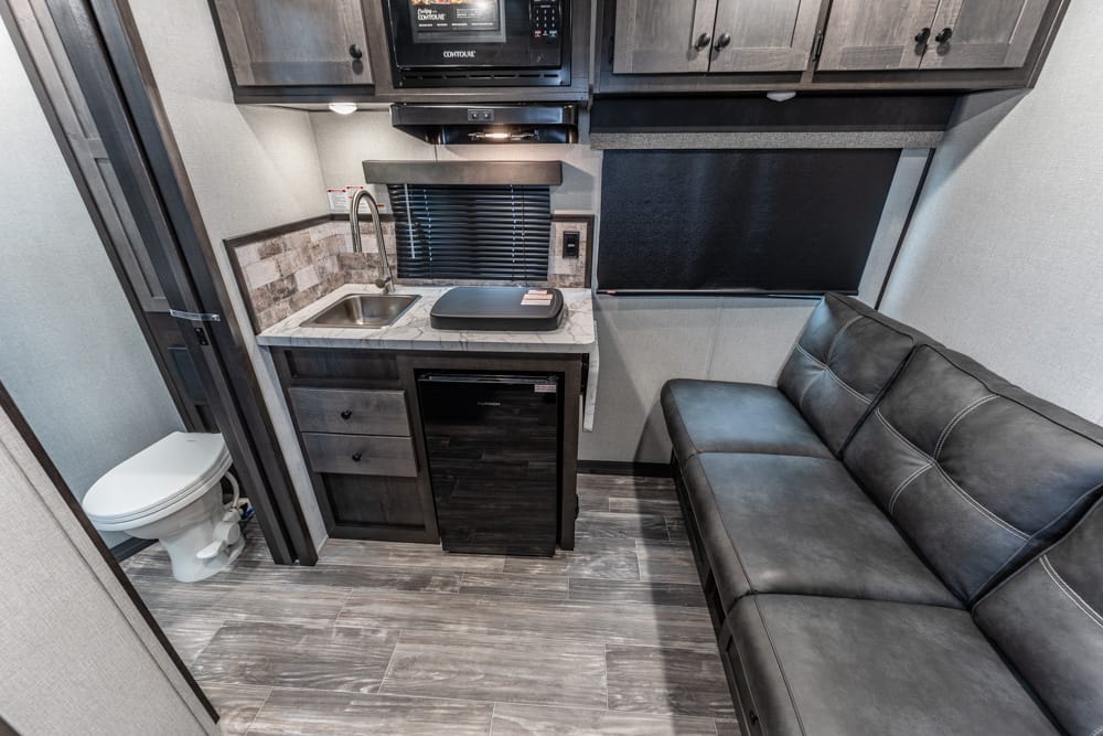 Compact RV kitchen with sink, small fridge, microwave, and a black leather sofa beside a window.
