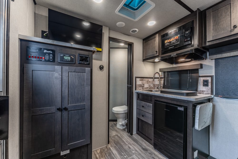 Interior of a modern RV kitchen with dark wood cabinets, stainless appliances, and a small fridge next to a sink and microwave.