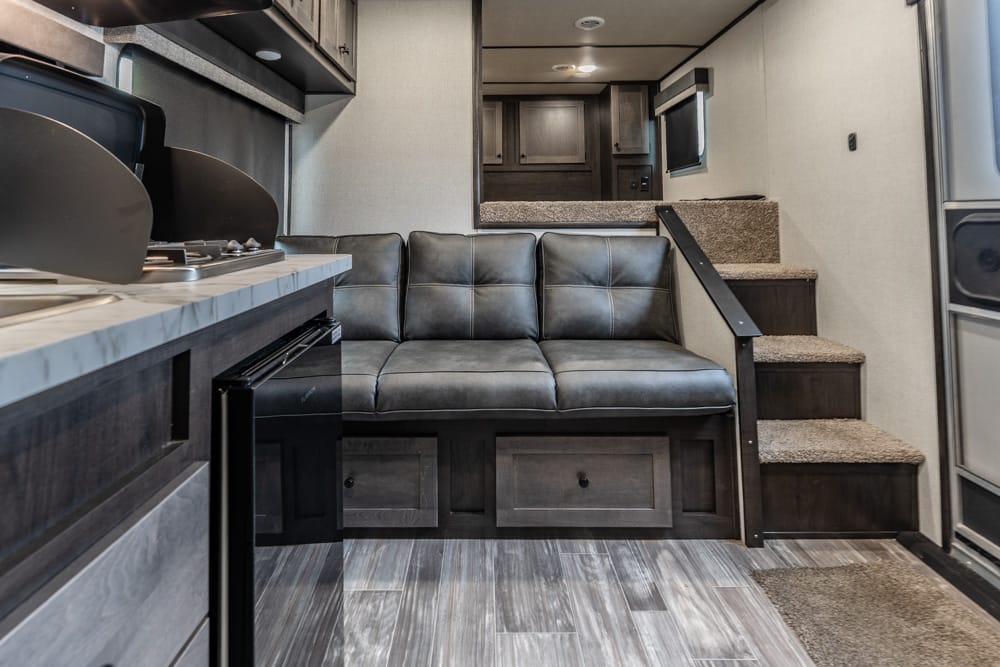 Interior of a modern RV living area: gray leather sofa, kitchen counter with stove on the left, dark wood cabinets, and carpeted stairs leading to an upper level.