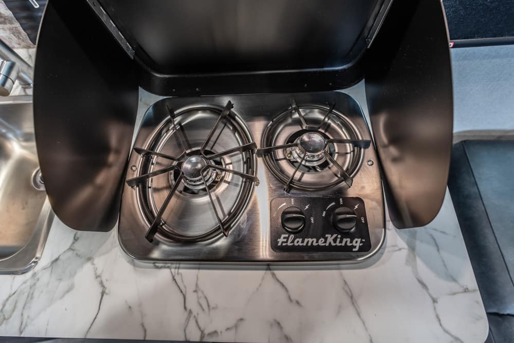 Two-burner stainless steel propane stove with open lid and FlameKing control dials on a marble countertop now ready for use.