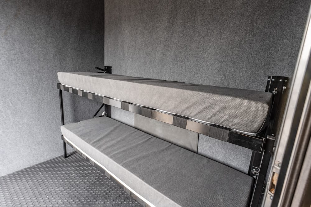 Two-tier bunk bed with gray cushions mounted to a black metal frame inside a small gray-walled space.