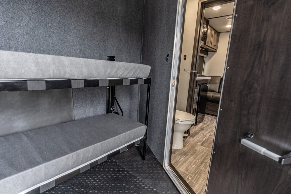 Interior of a compact trailer: dual bunk beds along the left wall and a doorway to the bathroom with a toilet visible.