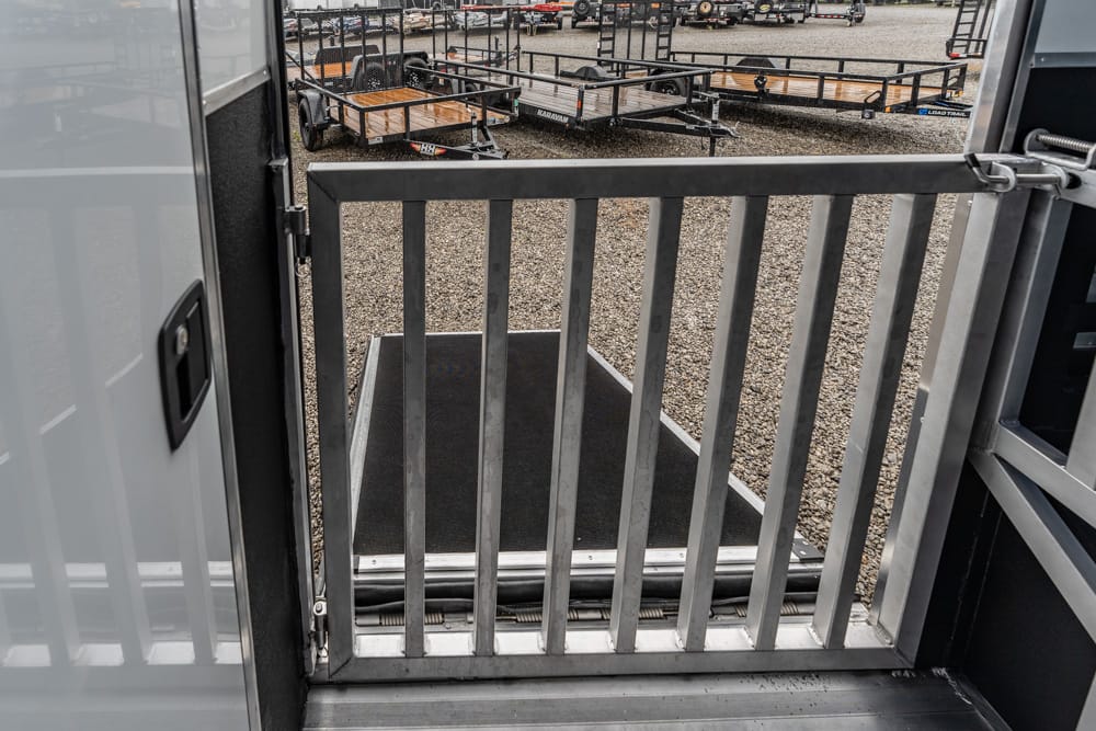 Interior view from inside a utility trailer, looking out through a vertical-bar metal ramp gate to a gravel lot with flatbed trailers in the background
