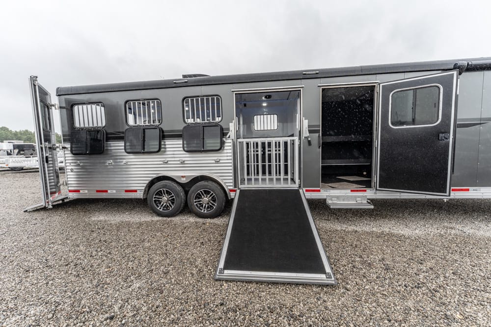 Horse trailer on a gravel lot with its rear ramp lowered for loading. The sides have windows and ventilation grilles, with a dark interior.