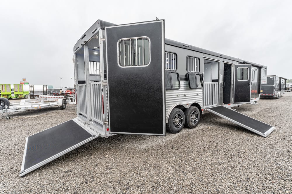 Black tandem-axle horse trailer with rear ramp lowered for loading on a gravel lot.