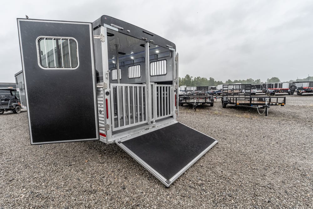 Rear view of a black horse trailer with the back doors open and a lowered ramp, parked on a gravel lot with other trailers in the background.