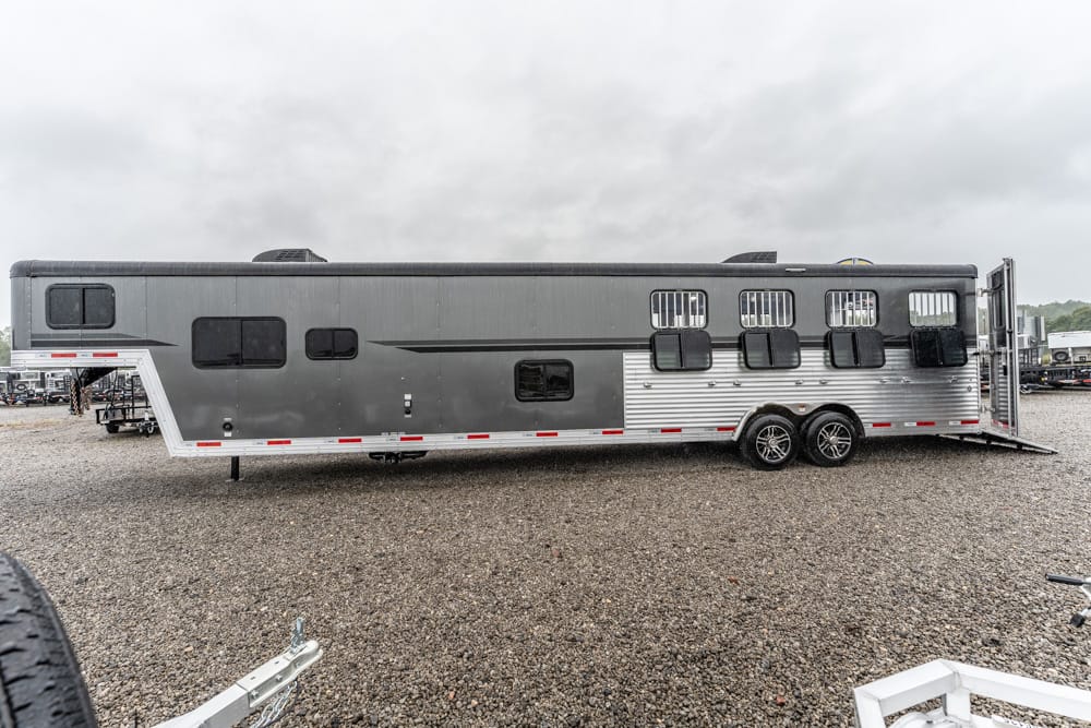 Long gray horse trailer with two axles parked on a gravel lot under a cloudy sky, rear loading ramp lowered.