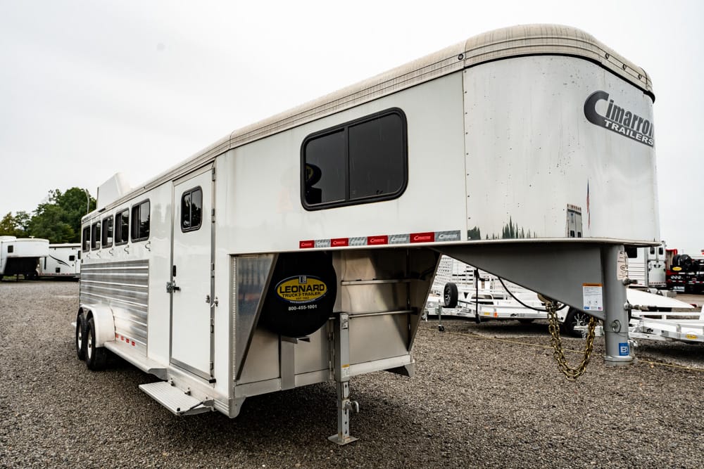 White Cimarron gooseneck horse trailer on a gravel lot, with a Leonard spare tire mounted near the hitch and chains visible at the hitch area.