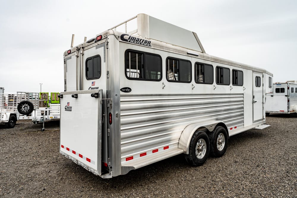 Silver two-axle horse trailer with a rear ramp and multiple side windows parked on a gravel lot; Cimarron Trailers branding visible on top edge of the roof.