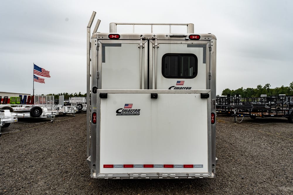 Rear view of a silver Cimarron trailer in a gravel lot, with a small window and American flags in the background.