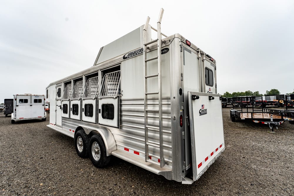 Silver horse trailer with barred stalls along the side, parked on a gravel lot with other trailers nearby
