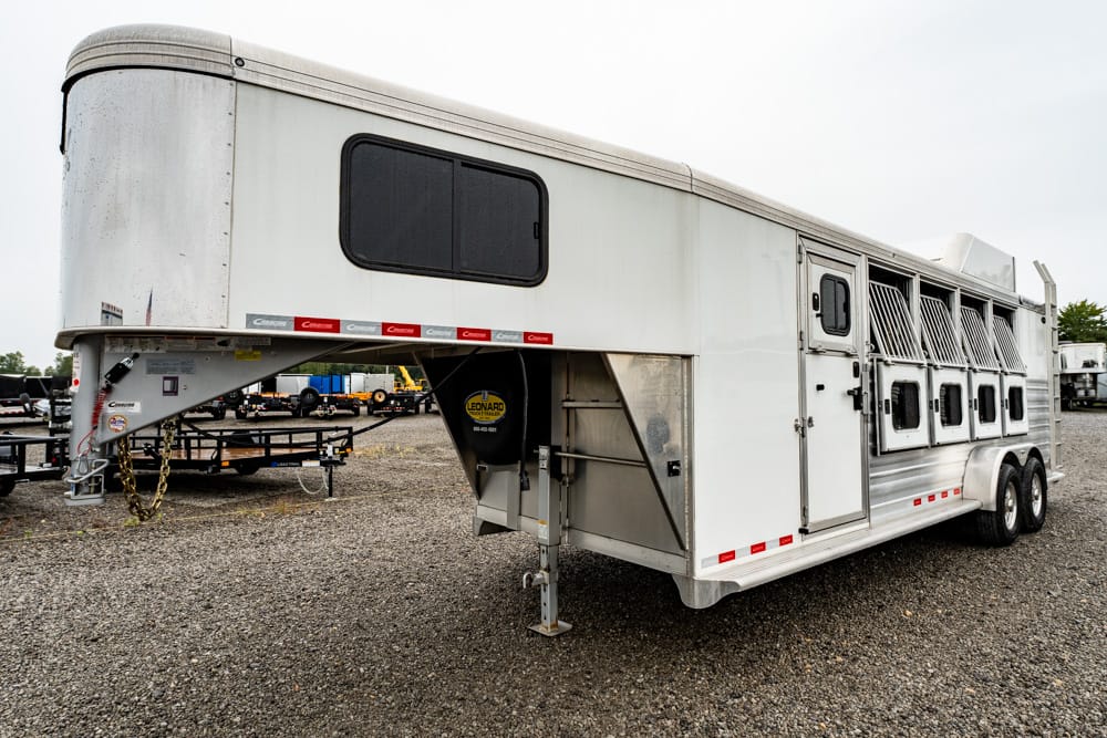 White gooseneck horse trailer with side ventilation stalls parked on a gravel lot, front hitch visible and a single window on the upper front section