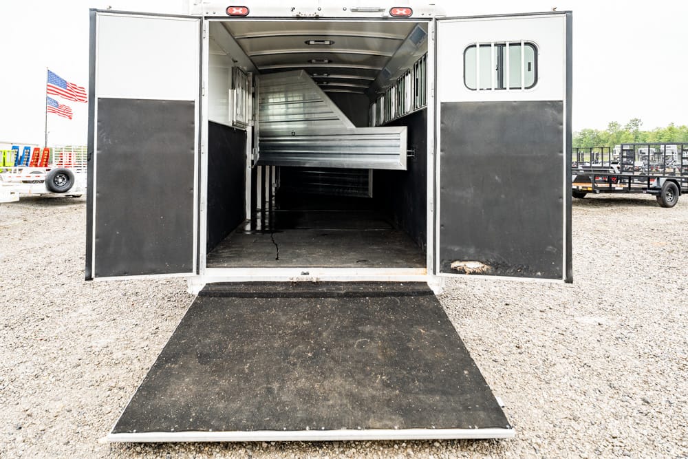 Open livestock/horse trailer with ramp lowered, showing a metal divider inside and a wide loading area on a gravel lot with other trailers in the background