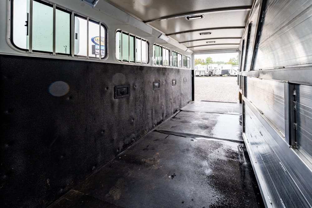 Interior of an empty livestock trailer with dark padded left wall, small windows along the top, and a wet floor; open rear shows a lot of parked trailers outside.