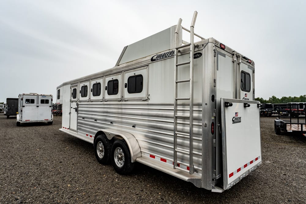 Silver aluminum horse trailer with multiple side windows and a roof ladder on a gravel lot.