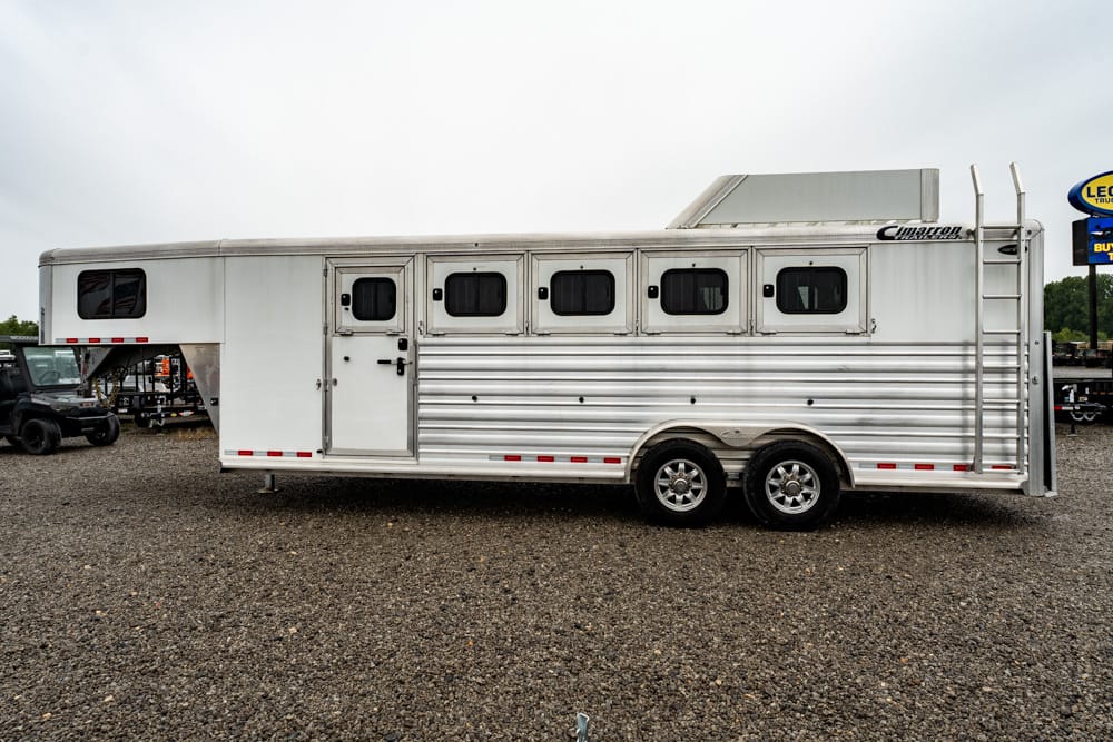 White horse trailer with multiple side windows, parked on a gravel lot; rear ladder attached to the back.