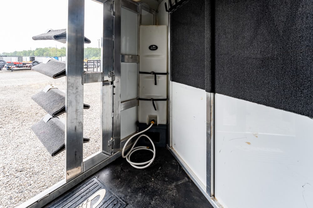 Interior of a trailer's utility area with a white stacked water tank, hoses on the floor, and metal framework nearby as the door is open to a gravel lot on the left.