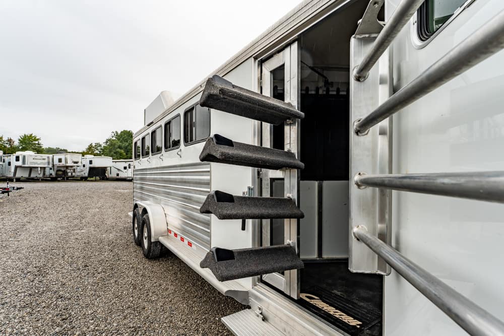 Open white horse trailer with folding black steps extended, interior stalls visible, parked on a gravel lot with other trailers in the background.