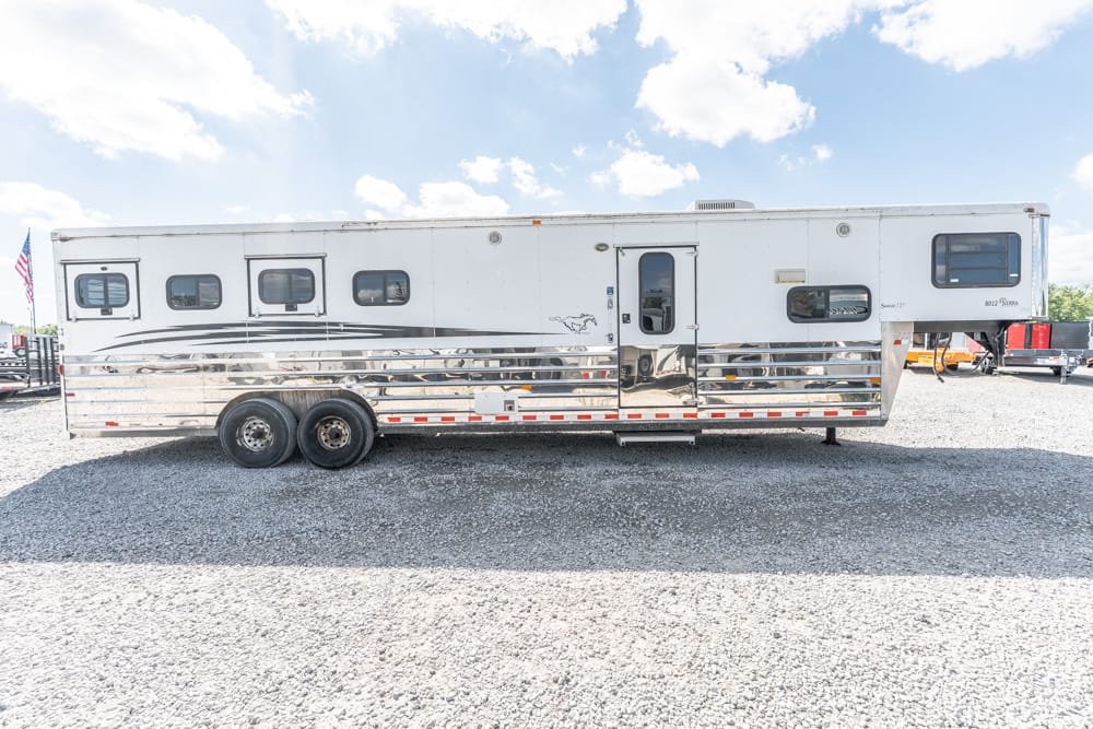 White aluminum horse trailer with chrome lower paneling on a gravel lot, two rear axles and small windows set along the side under a blue sky.