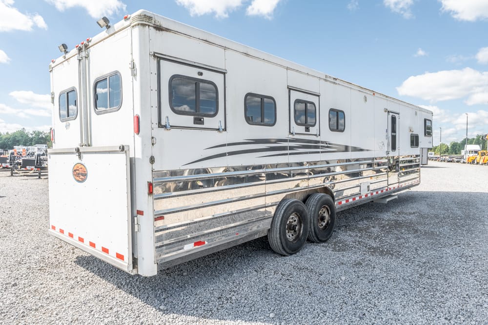 White aluminum horse trailer with multiple side windows and dual axles on a gravel lot, captured from a rear-side angle.