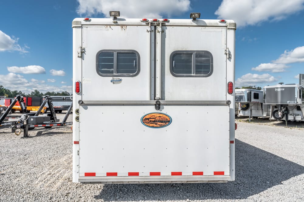 Rear view of a white trailer with two small windows, a badge emblem, and red taillights on a gravel lot.