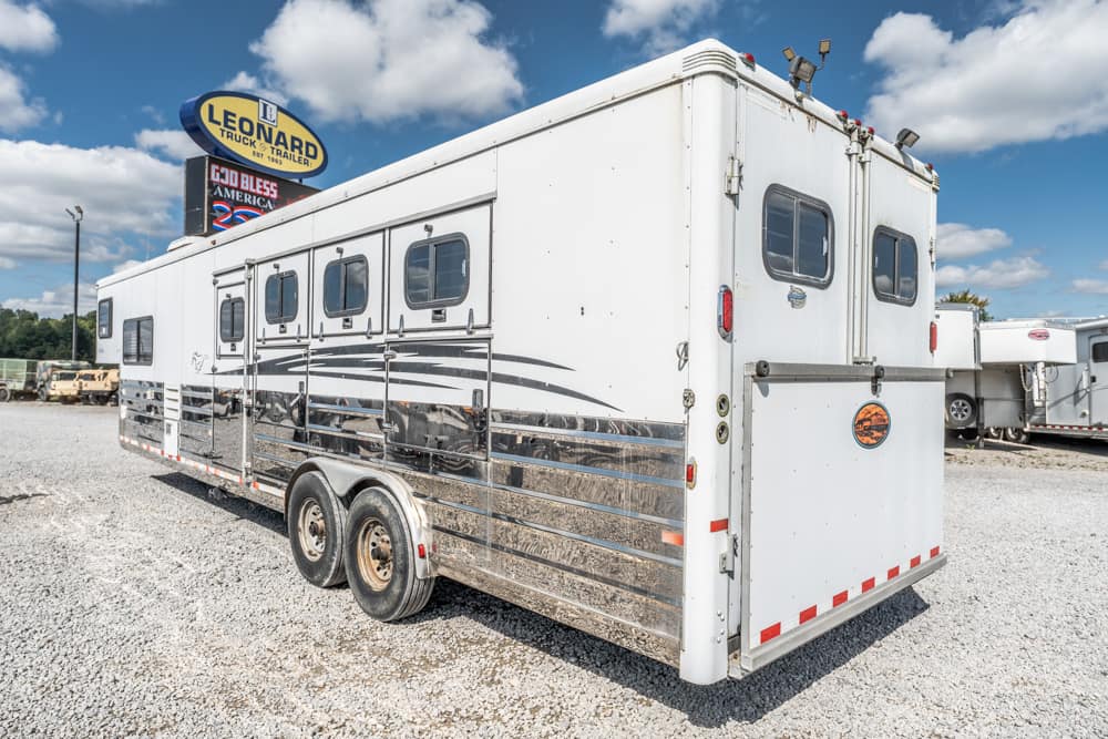 White aluminum horse trailer on a gravel lot with small windows and chrome lower siding, Leonard Truck & Trailer sign in the background.