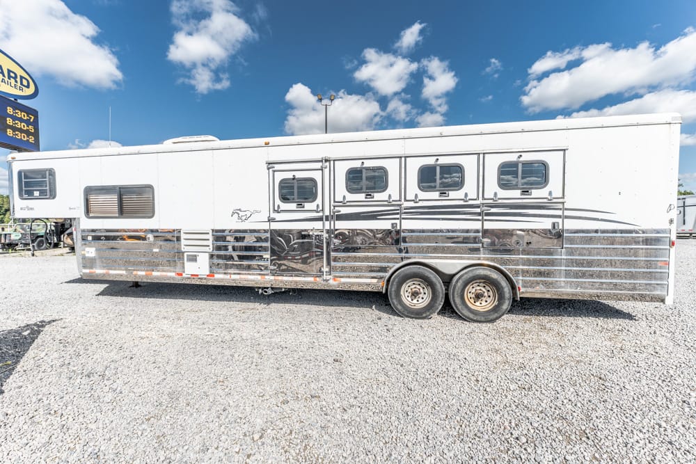 White aluminum horse trailer with six windows and chrome lower panels, parked on a gravel lot.