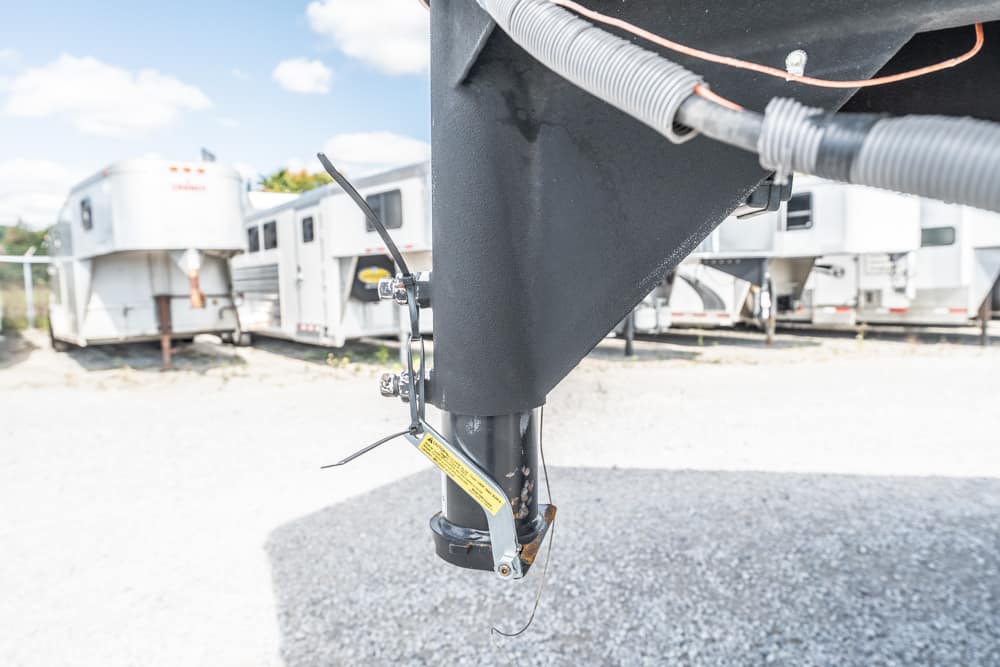 Close-up of a trailer hitch with a metal jack, safety chains, and a yellow warning label on a gravel lot.