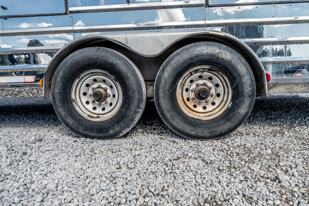 Twin trailer wheels with chrome rims on a gravel surface beside a chrome-trimmed trailer.