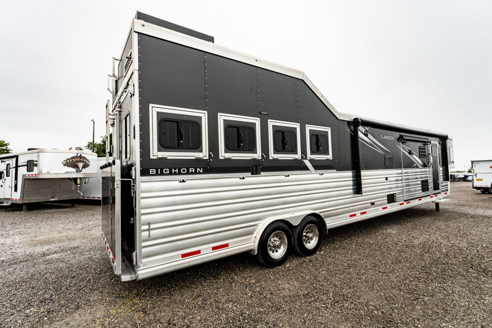 Long two-axle horse trailer with black upper panels and silver corrugated lower siding parked on a gravel lot, showing multiple windows and a back ramp.