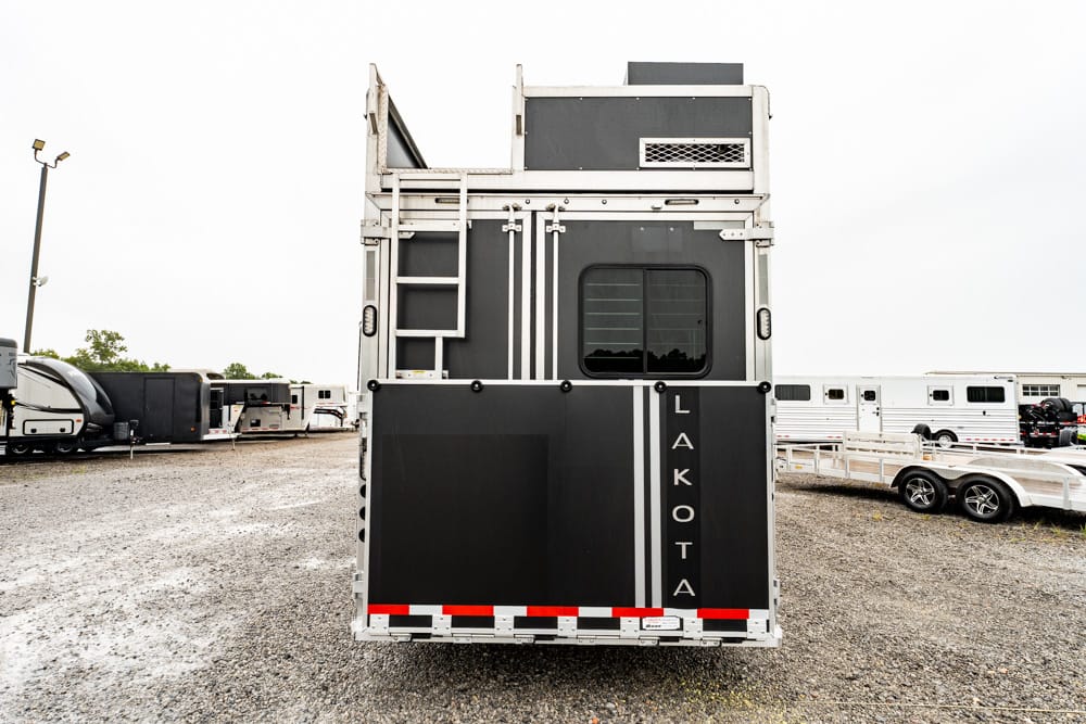 Rear view of a black and gray Lakota horse trailer with vertical letters, in a gravel lot among other trailers and equipment