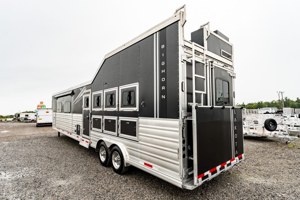Long silver and black horse trailer with multiple side windows and a rear ramp on a gravel lot
