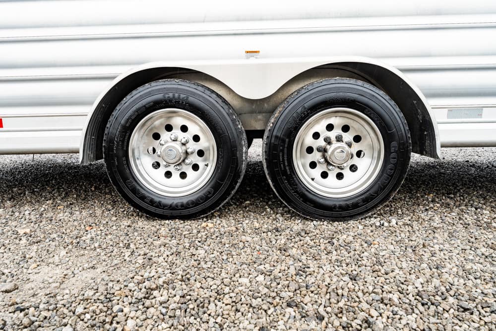 Dual trailer wheels with metal rims under a silver trailer on a gravel surface, showing the wheel wells and tires