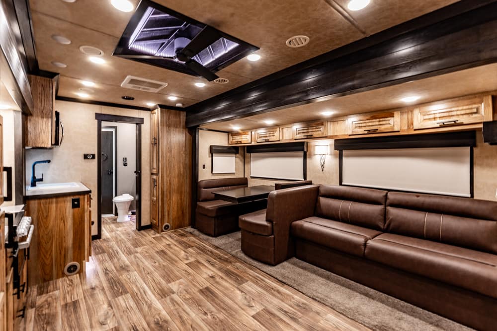 Interior of a luxury RV living area with brown leather sofas, a booth dining area, and overhead cabinets, plus wood-like flooring and warm lighting.
