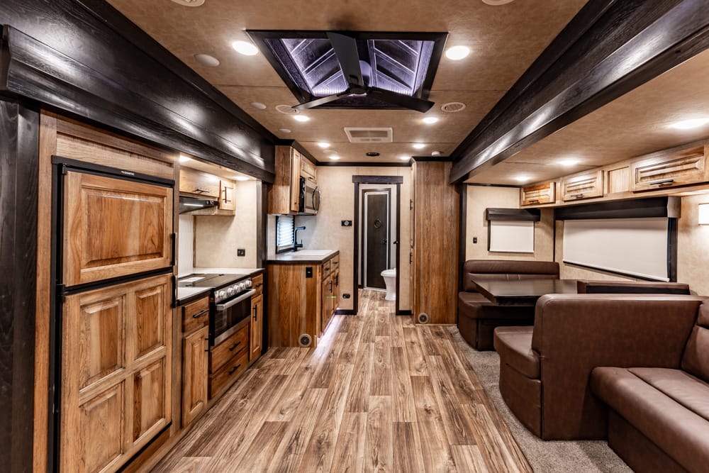Interior of a modern recreational vehicle: wood cabinets, full kitchen on the left, and brown leather seating with a dinette on the right. Bathroom at the far end.