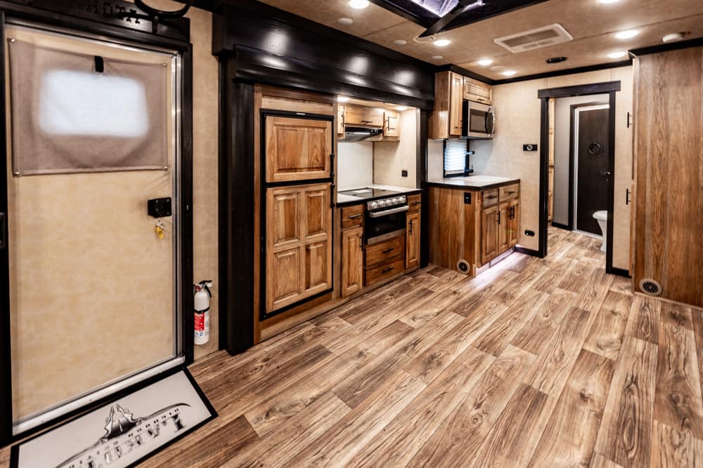Interior of a wood-finished RV kitchen with cabinets, stove, oven, and a hallway leading to a bathroom. Wooden plank flooring throughout.
