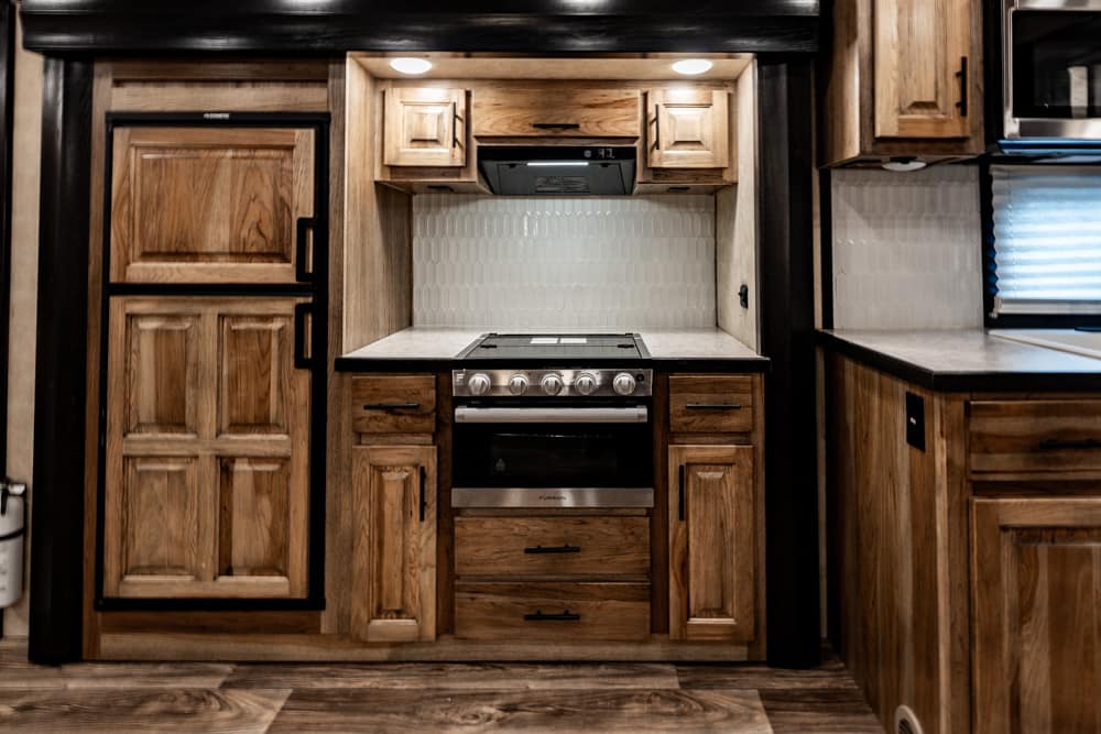 Wooden RV kitchen with a stainless steel oven and four-burner stove, tile backsplash, and upper cabinets surrounding a compact workspace.",