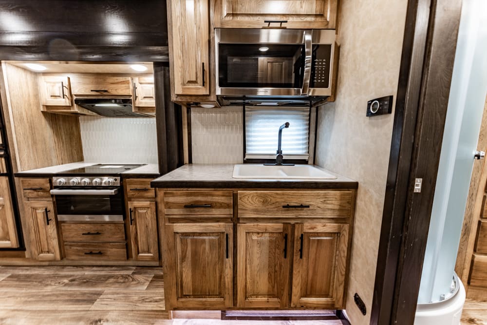 Wooden RV kitchen with stove, oven, microwave, and double sink under a window.