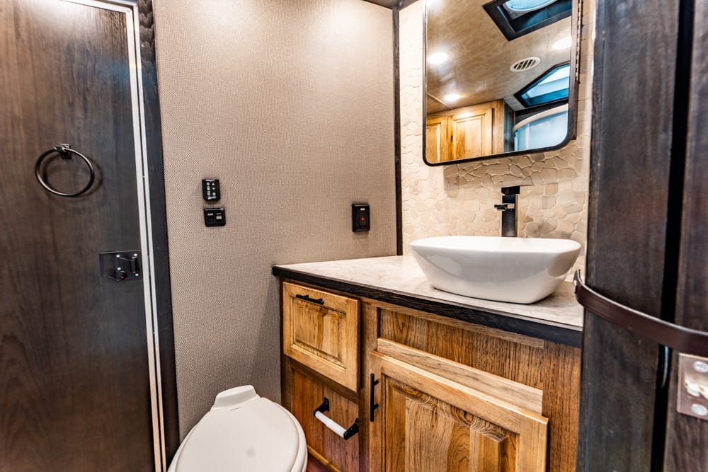RV bathroom interior with a vessel sink on a marble countertop, wooden vanity, and oval mirror above a pebble-textured wall. Windows and ceiling light reflections visible.